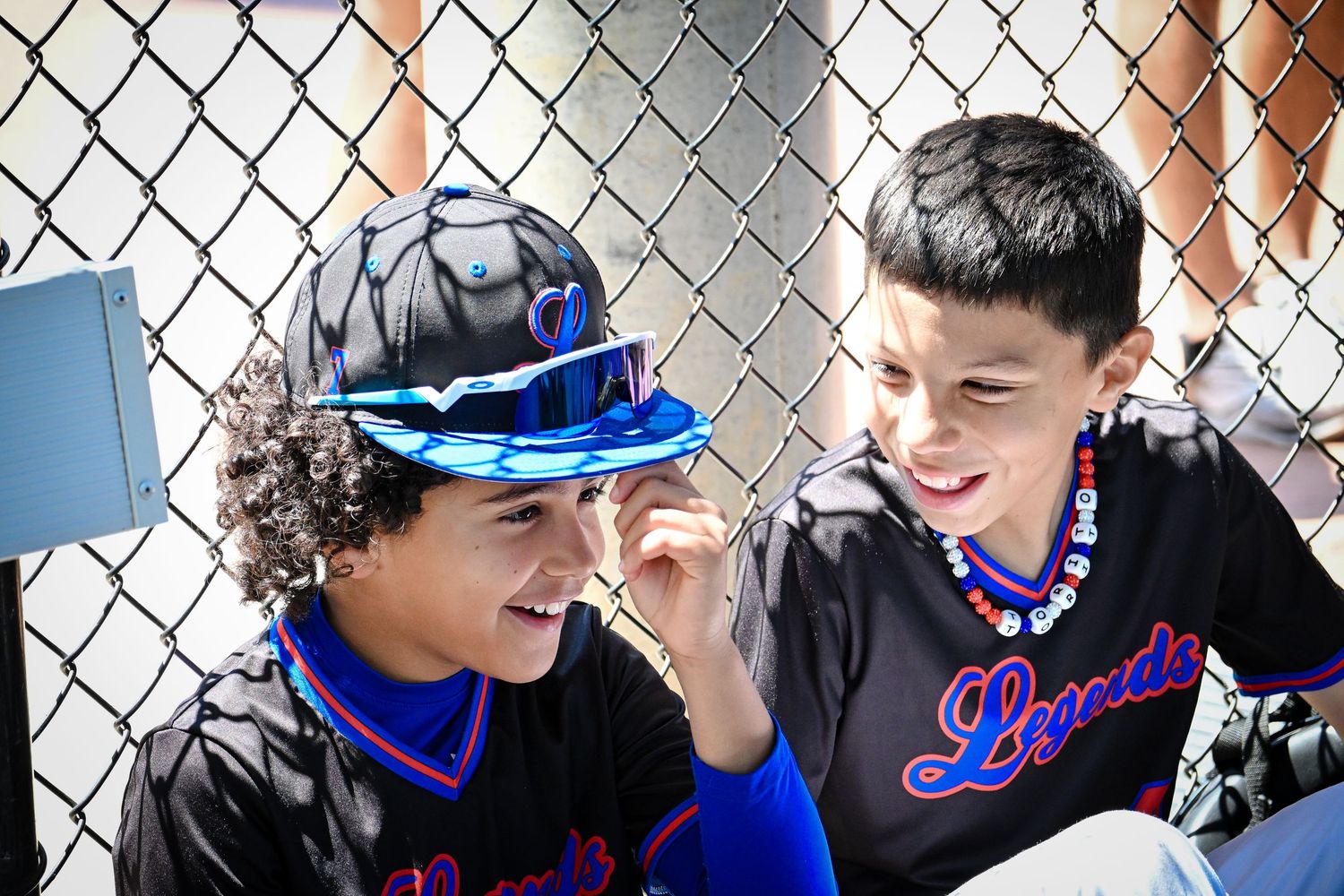 Two young baseball players in Lucado team jerseys laugh together near a chain-link fence.