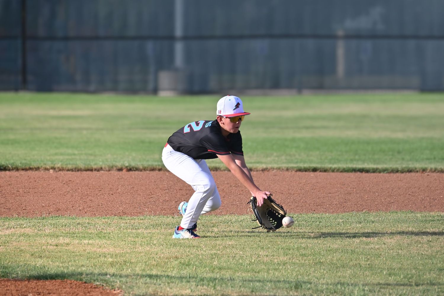 Young baseball player in white uniform crouching in ready position to field ball on sunny infield.