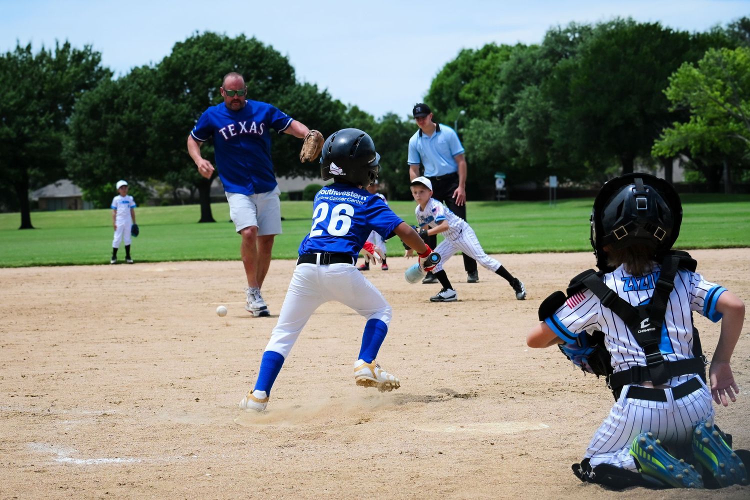 Youth baseball batter in Rangers uniform #26 swings at pitch with catcher and coach nearby.