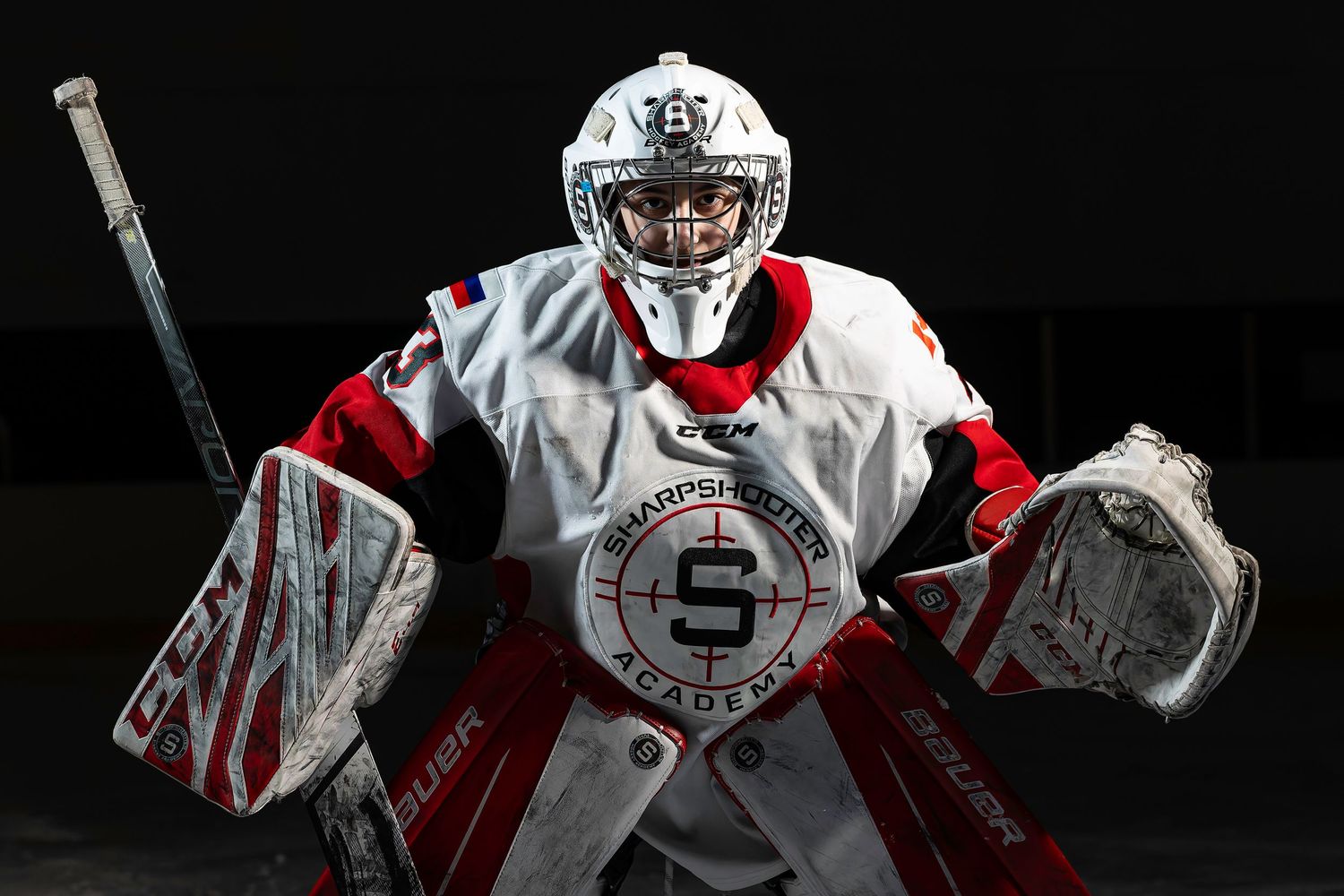 Ice hockey goalie in white Shattuck-Saint Mary's Academy uniform crouches in ready position on dark rink.