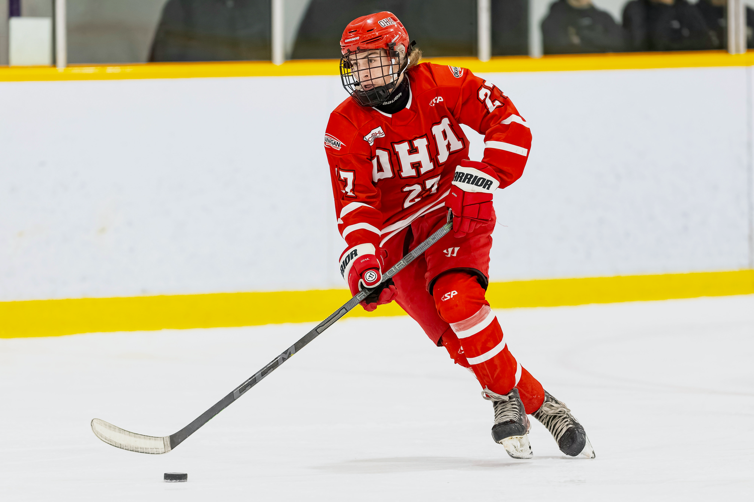 Hockey player in red OHA jersey number 27 skating with puck on indoor ice rink.