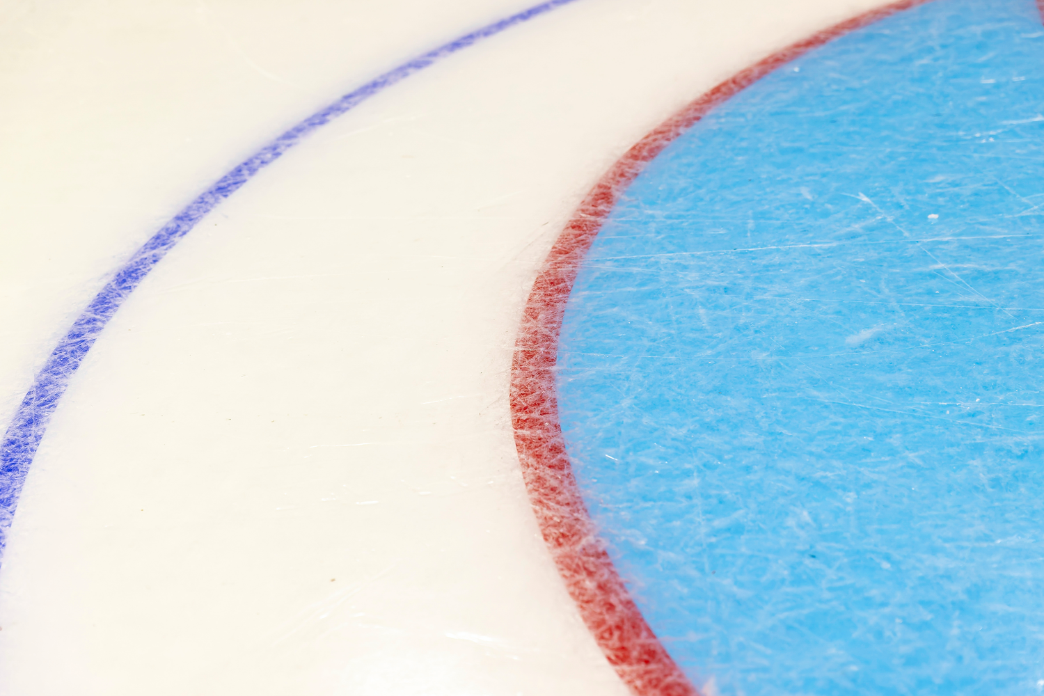 Close-up of a hockey rink ice surface featuring a blue face-off circle with red border and a blue line marking.