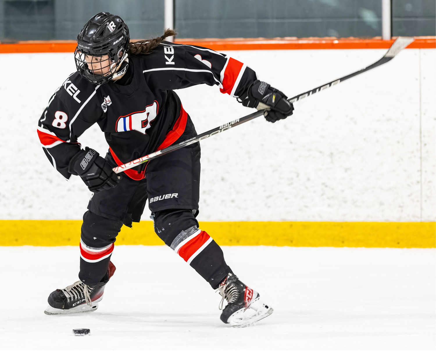 Youth hockey player in black jersey number 8 skating and shooting puck on indoor ice rink.