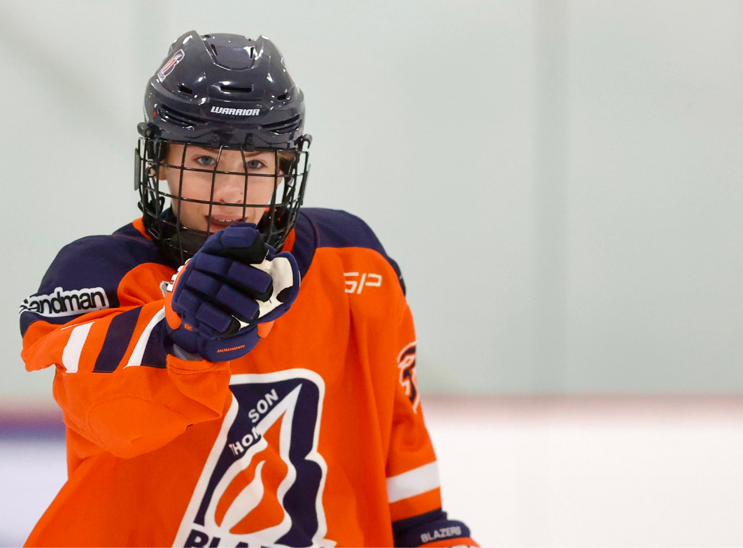 Hockey player in orange jersey and helmet gestures on ice rink during game.