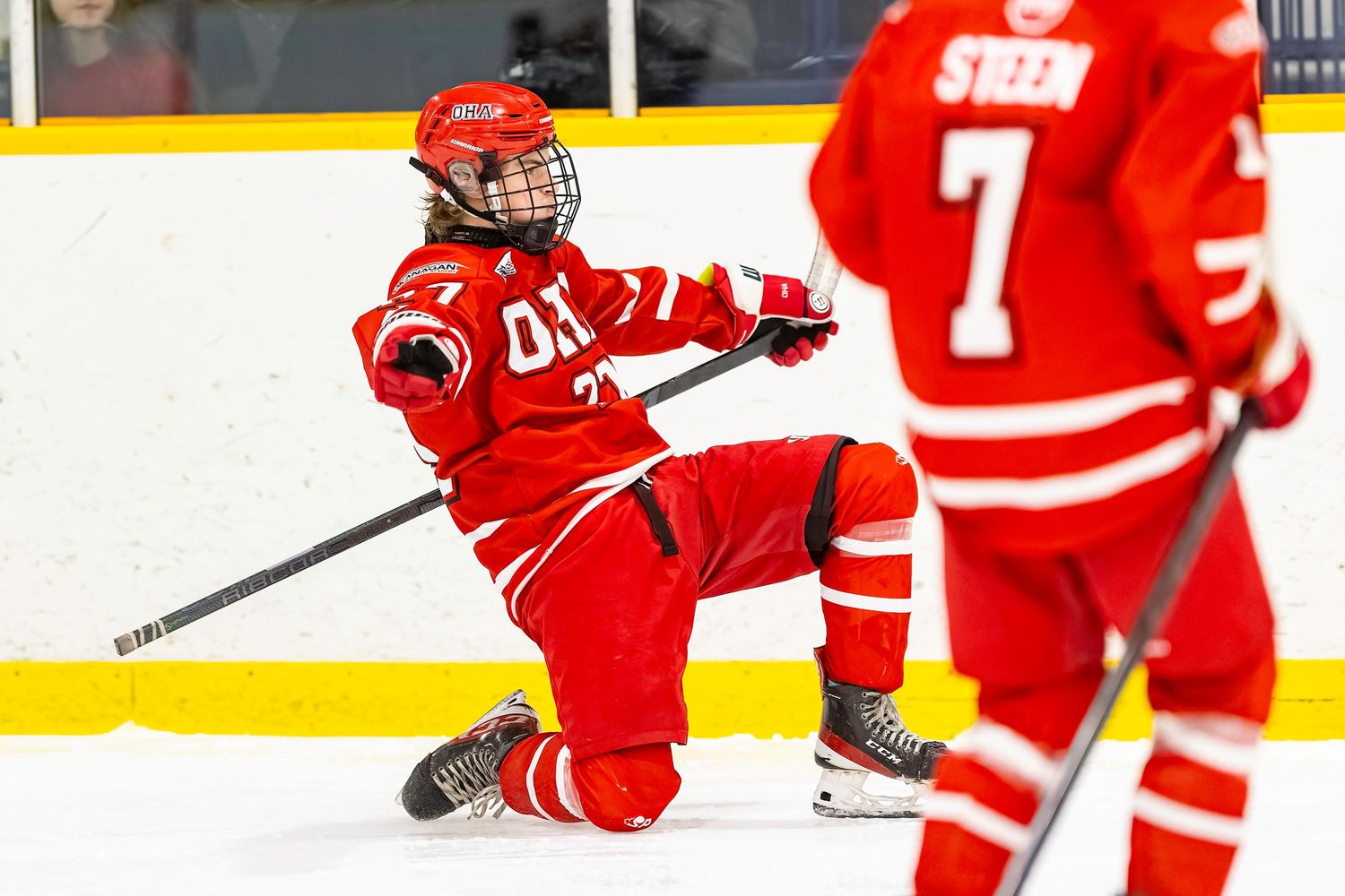 Hockey player in red uniform celebrating on ice, sliding on knees with stick raised during a game.