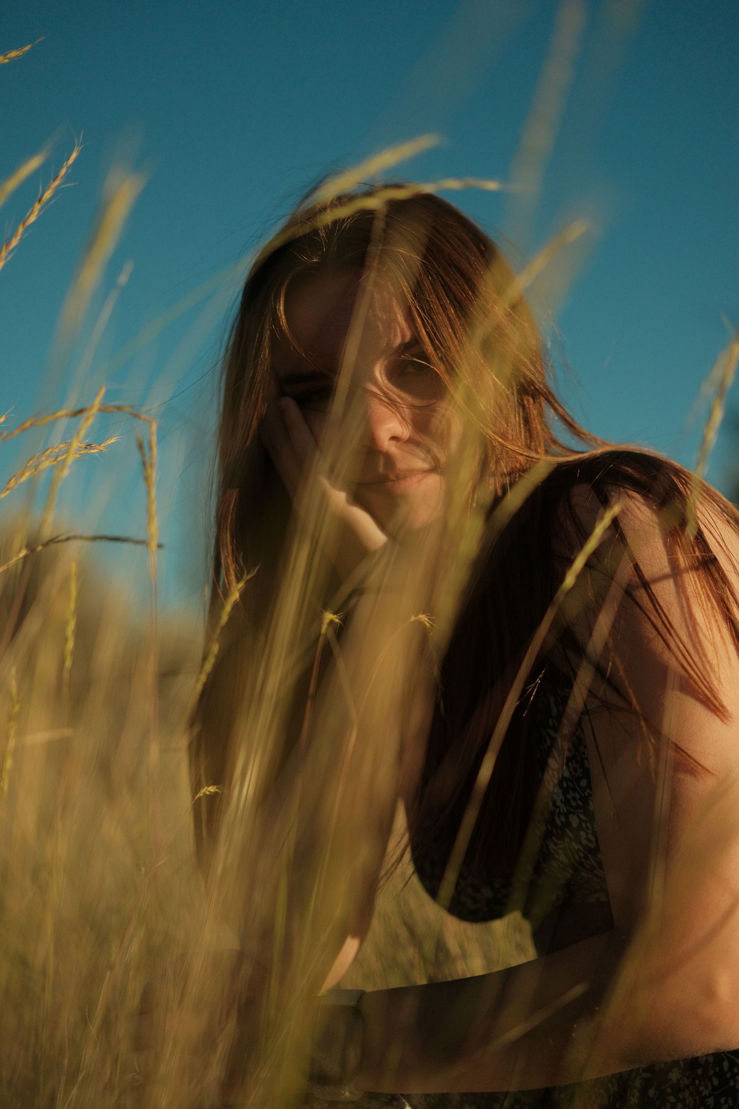 Young woman with long hair peers through tall golden grass blades under a vivid blue sky.