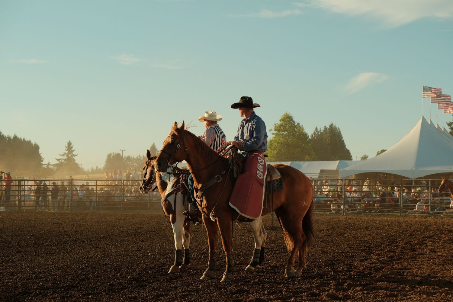 Two cowboys on horseback in a rodeo arena during golden hour with spectators in the background.