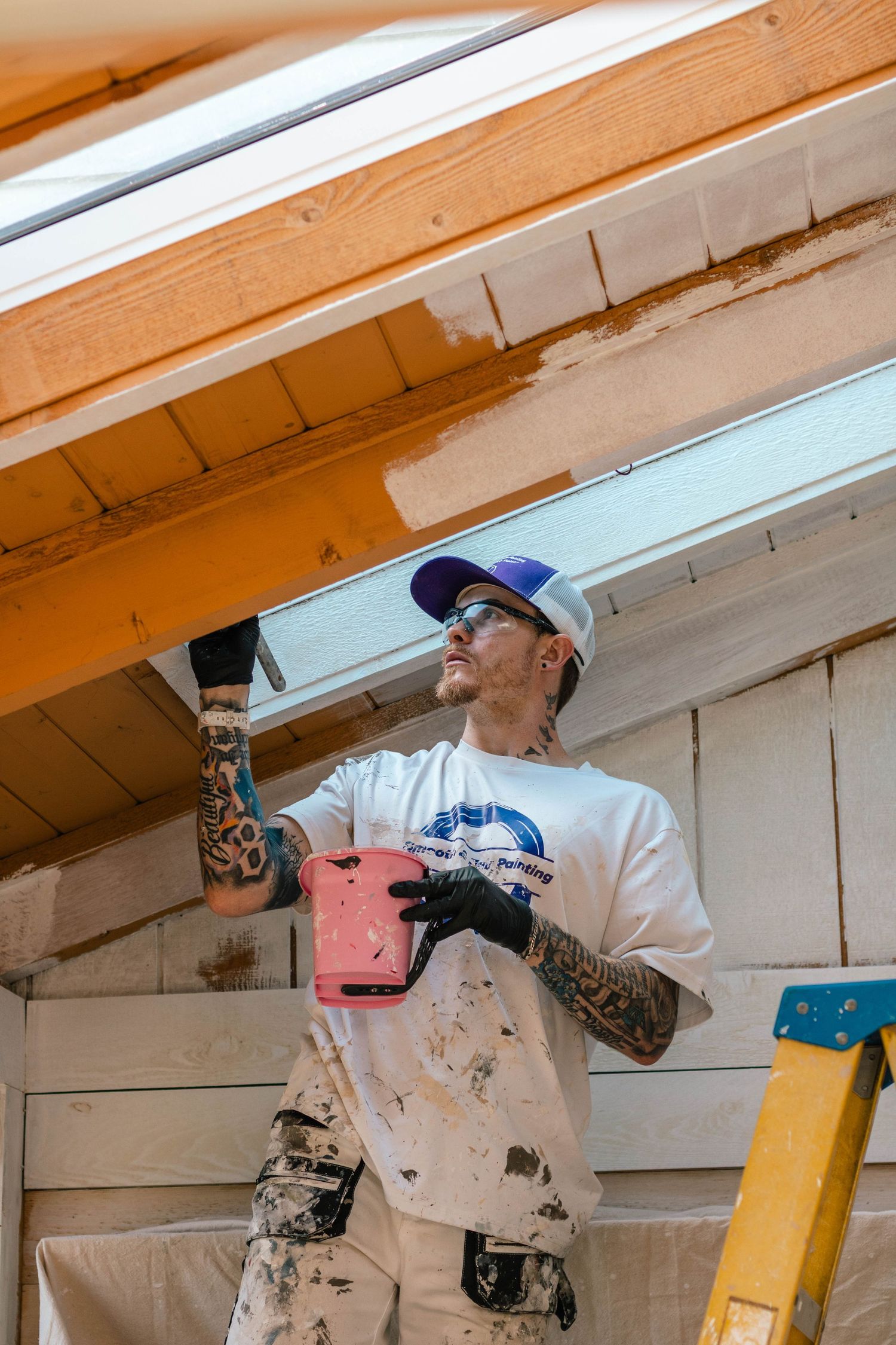 Tattooed male painter in white shirt and cap carefully painting wooden ceiling beams on a ladder.