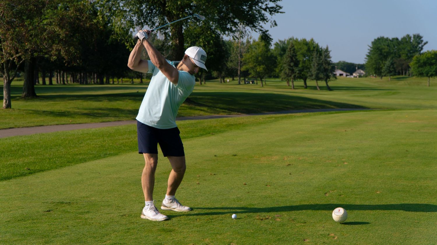 Golfer in white shirt and cap swings a driver on a lush green golf course surrounded by trees.