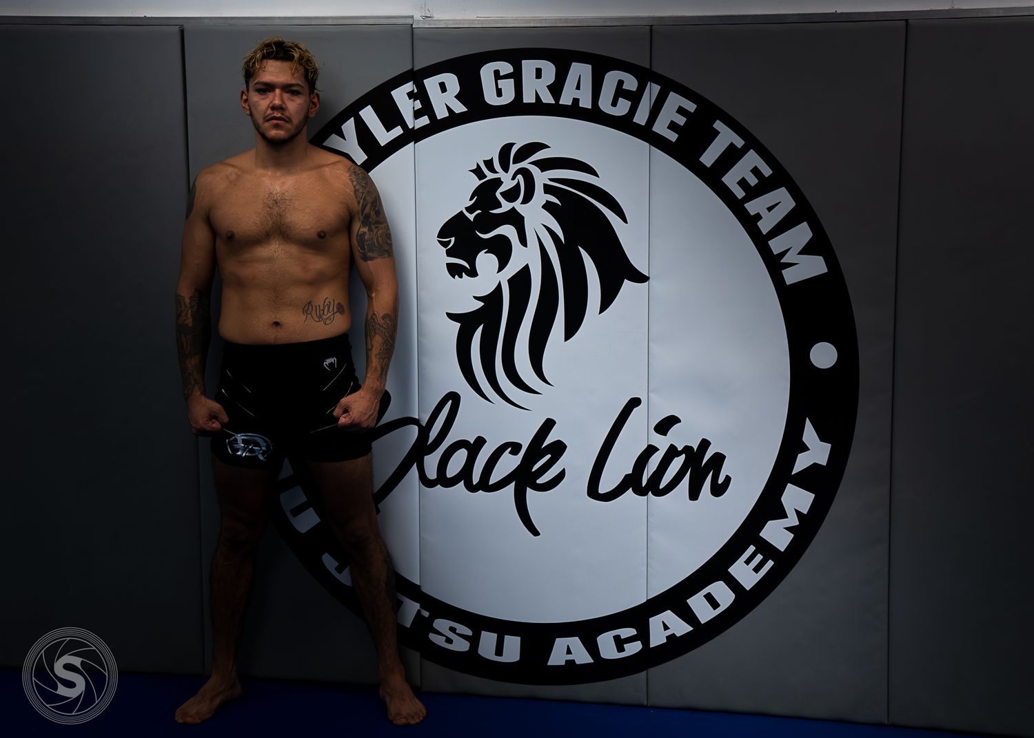 Shirtless male MMA fighter poses in front of Black Lion Jiu Jitsu Academy sign at Yler Gracie Team gym.