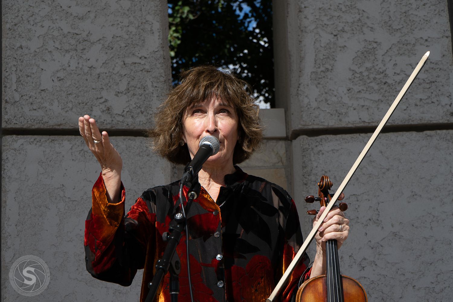 Woman playing violin and speaking into microphone outdoors at a live music performance event.