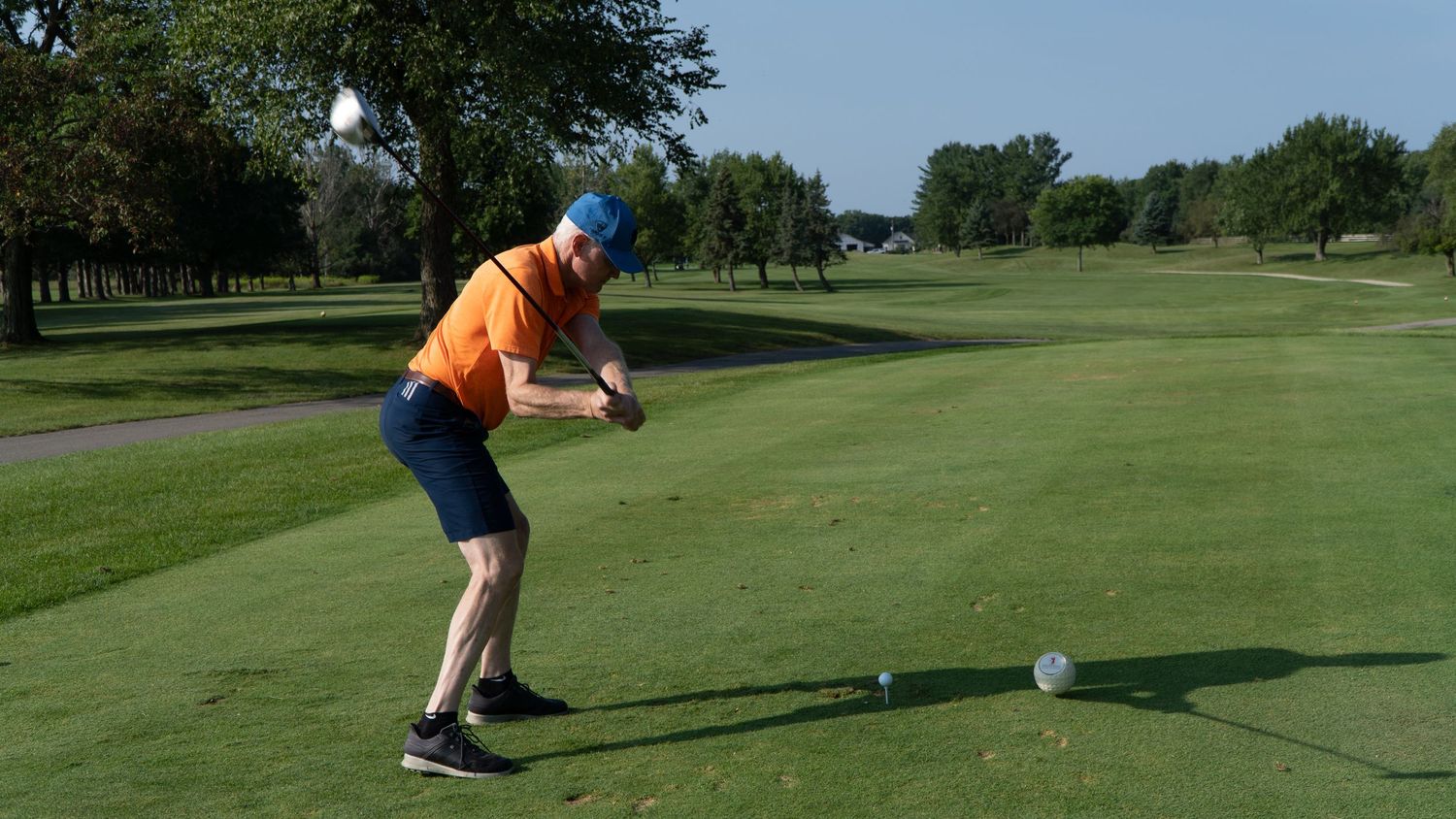 Golfer in orange shirt and blue hat swinging a club on a lush green golf course on a sunny day.