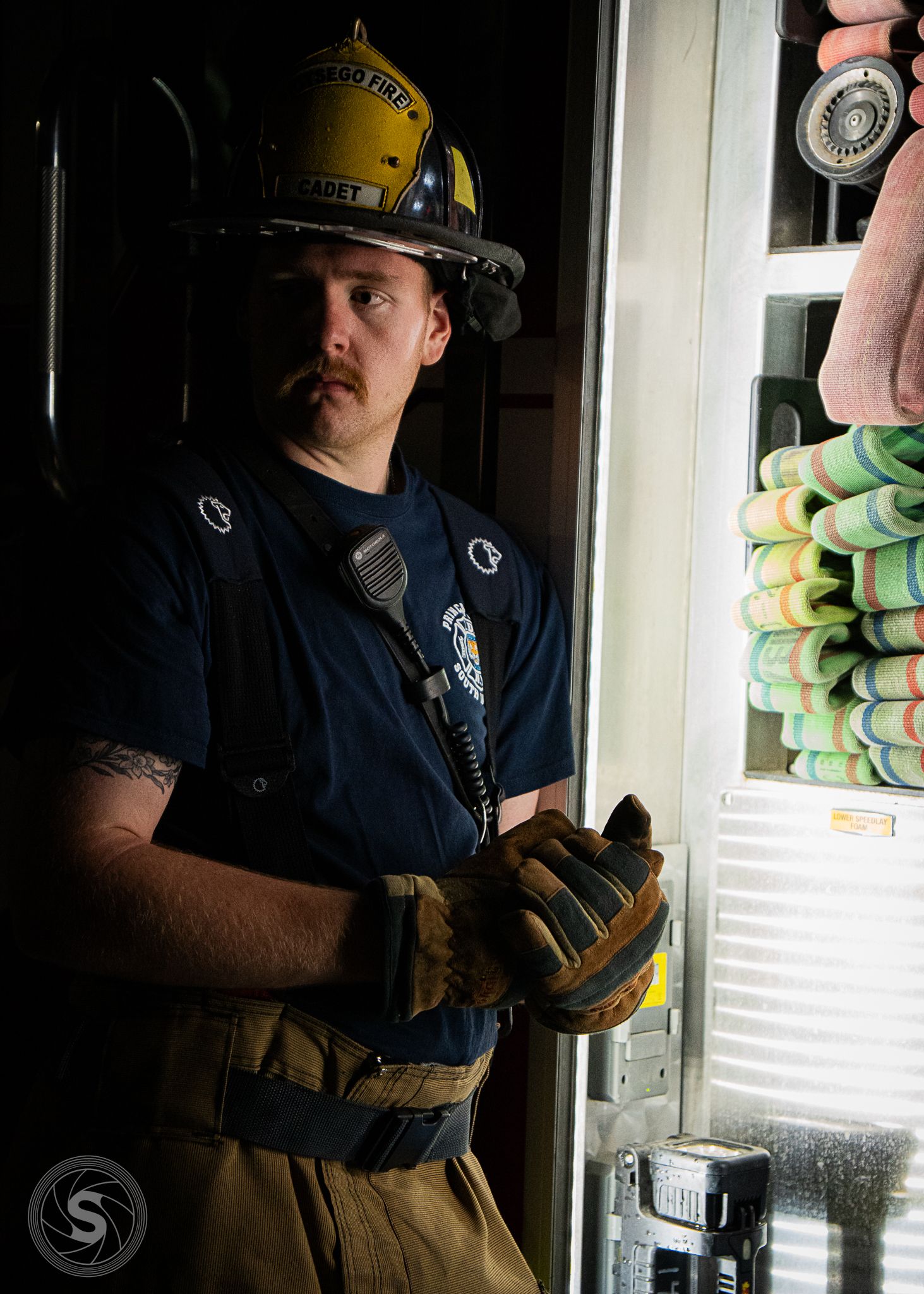 A firefighter in yellow helmet and tactical gloves stands by a fire truck, wearing a navy uniform with radio.