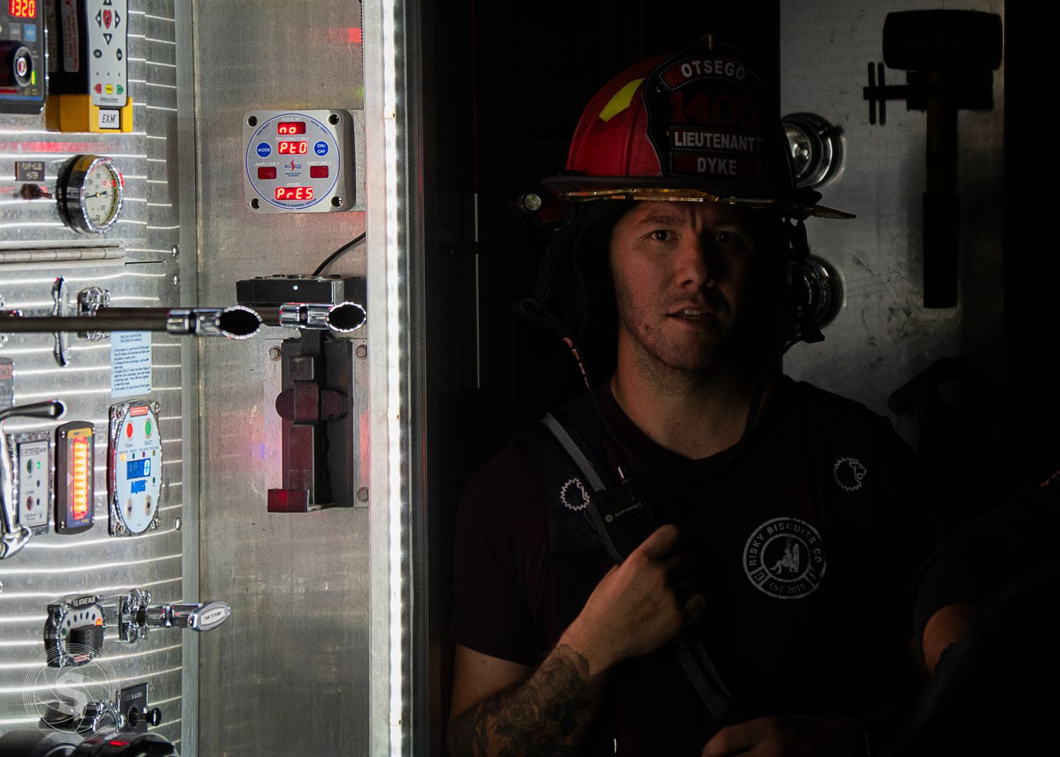 Firefighter in helmet and gear standing inside a fire station truck bay, holding equipment in dramatic lighting.