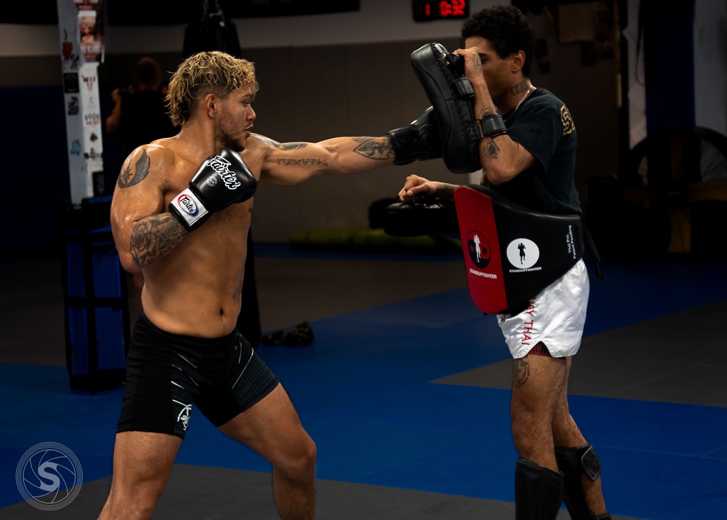 Two MMA fighters training with punch mitts in a gym, one shirtless fighter throwing a punch at a pad held by his partner.