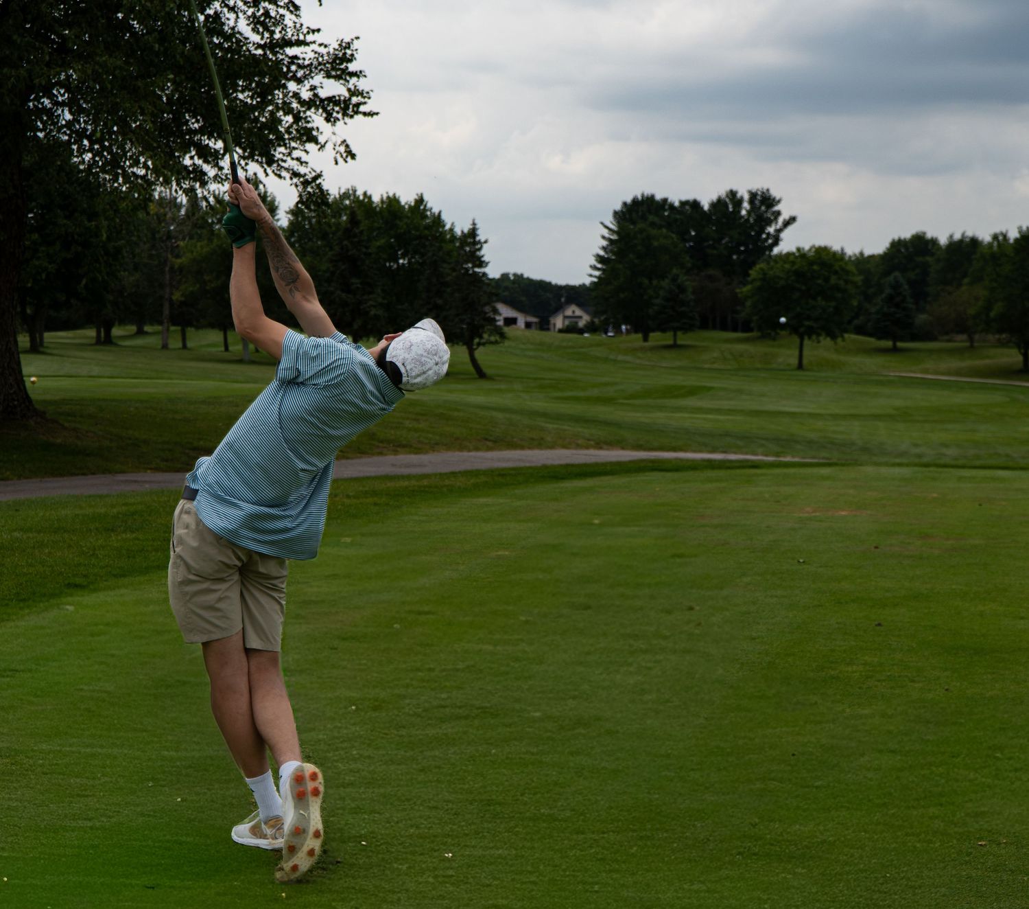 Golfer in blue shirt and khaki shorts swinging a golf club on a lush green golf course under cloudy skies.