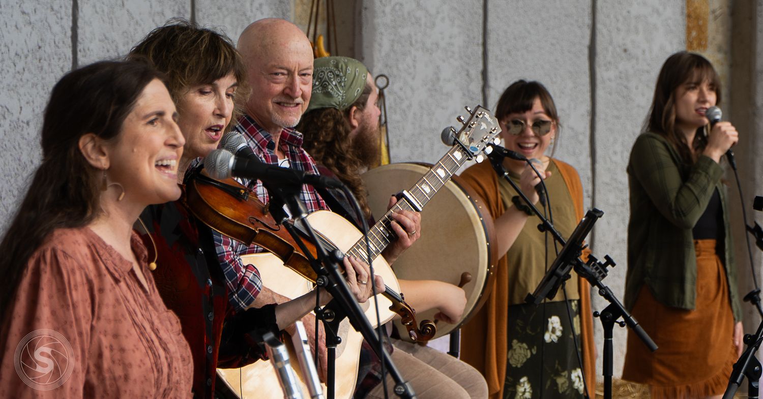 A folk band performs outdoors, featuring violin, guitar, and bodhran players singing together at microphones.