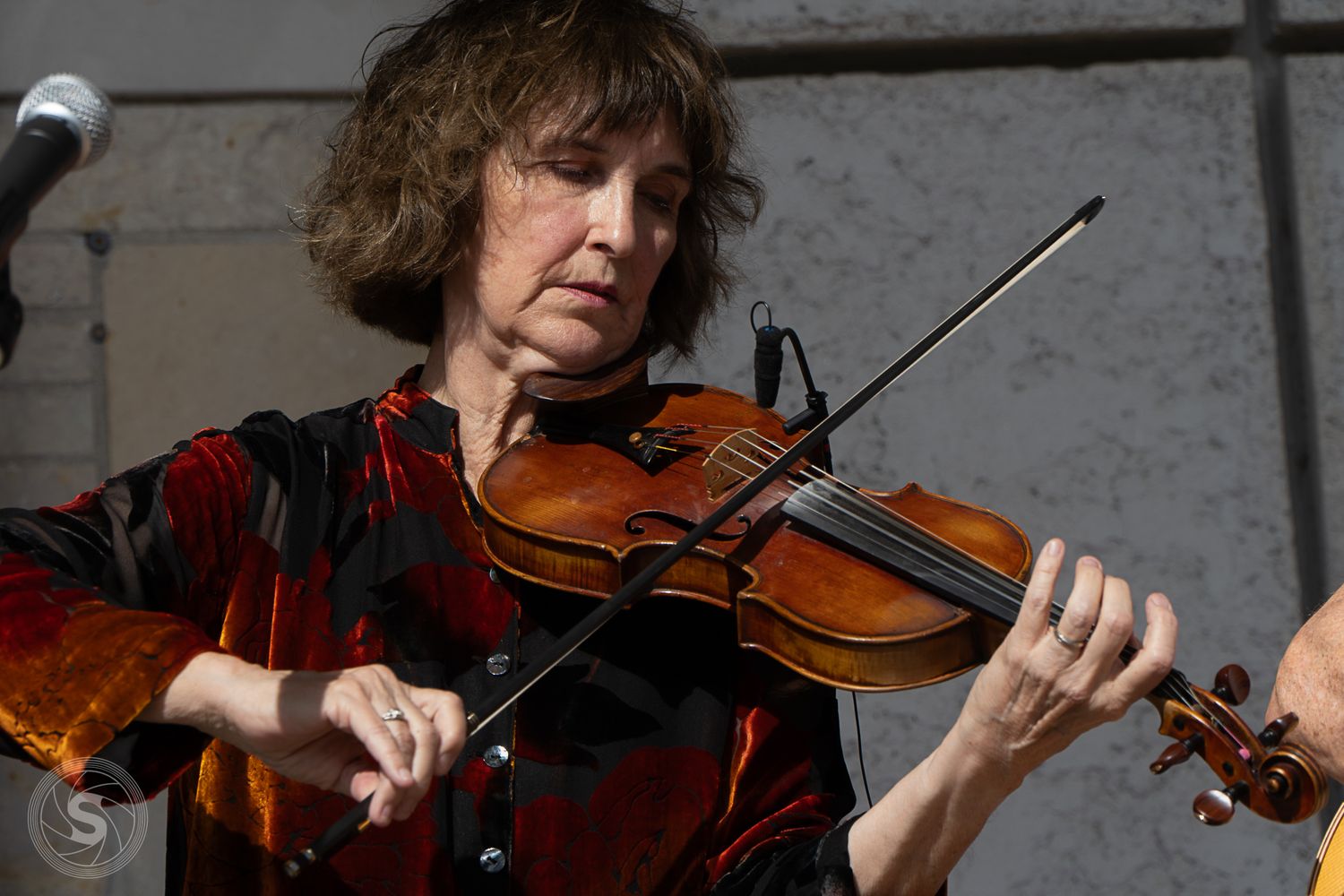 A woman with short brown hair plays violin outdoors, wearing a red patterned top during a live performance.