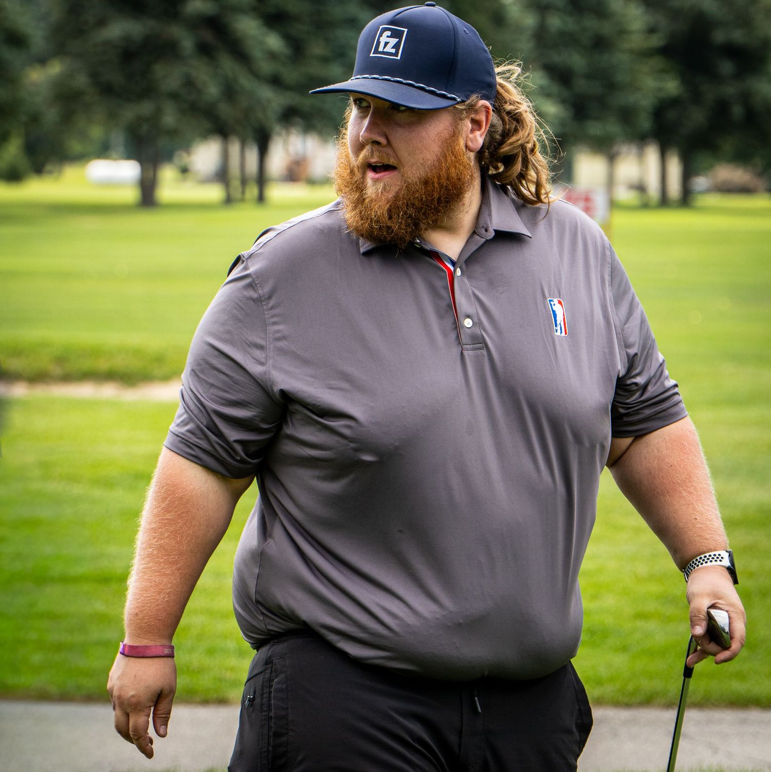 Bearded man in gray polo shirt and navy cap holding a golf club on a lush green golf course.