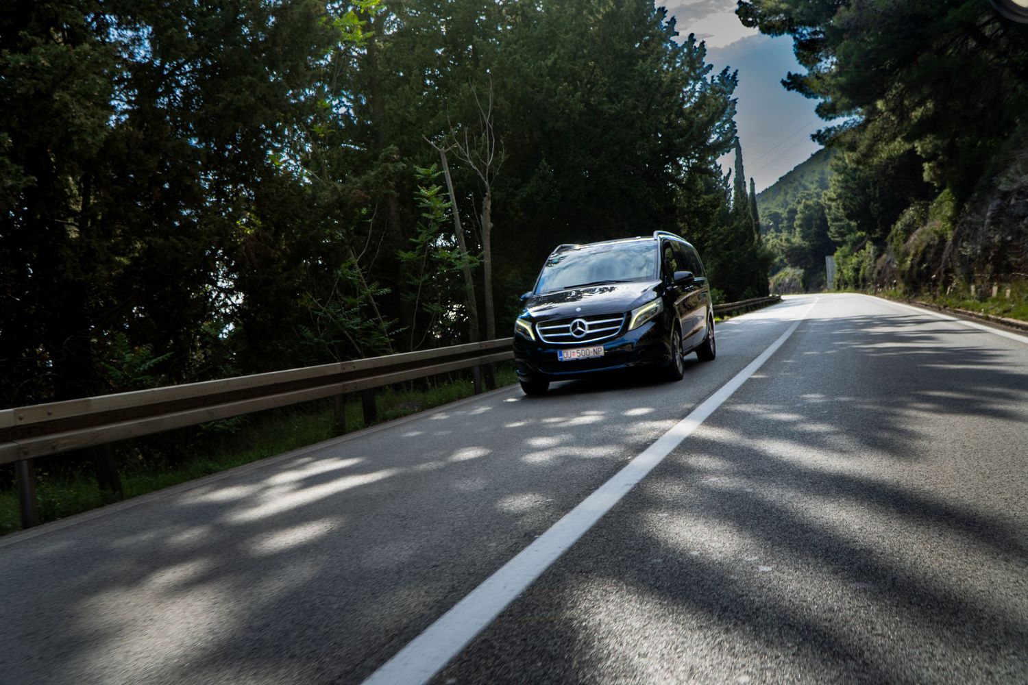 A black Mercedes-Benz van driving on a winding tree-lined road in a scenic mountainous area.
