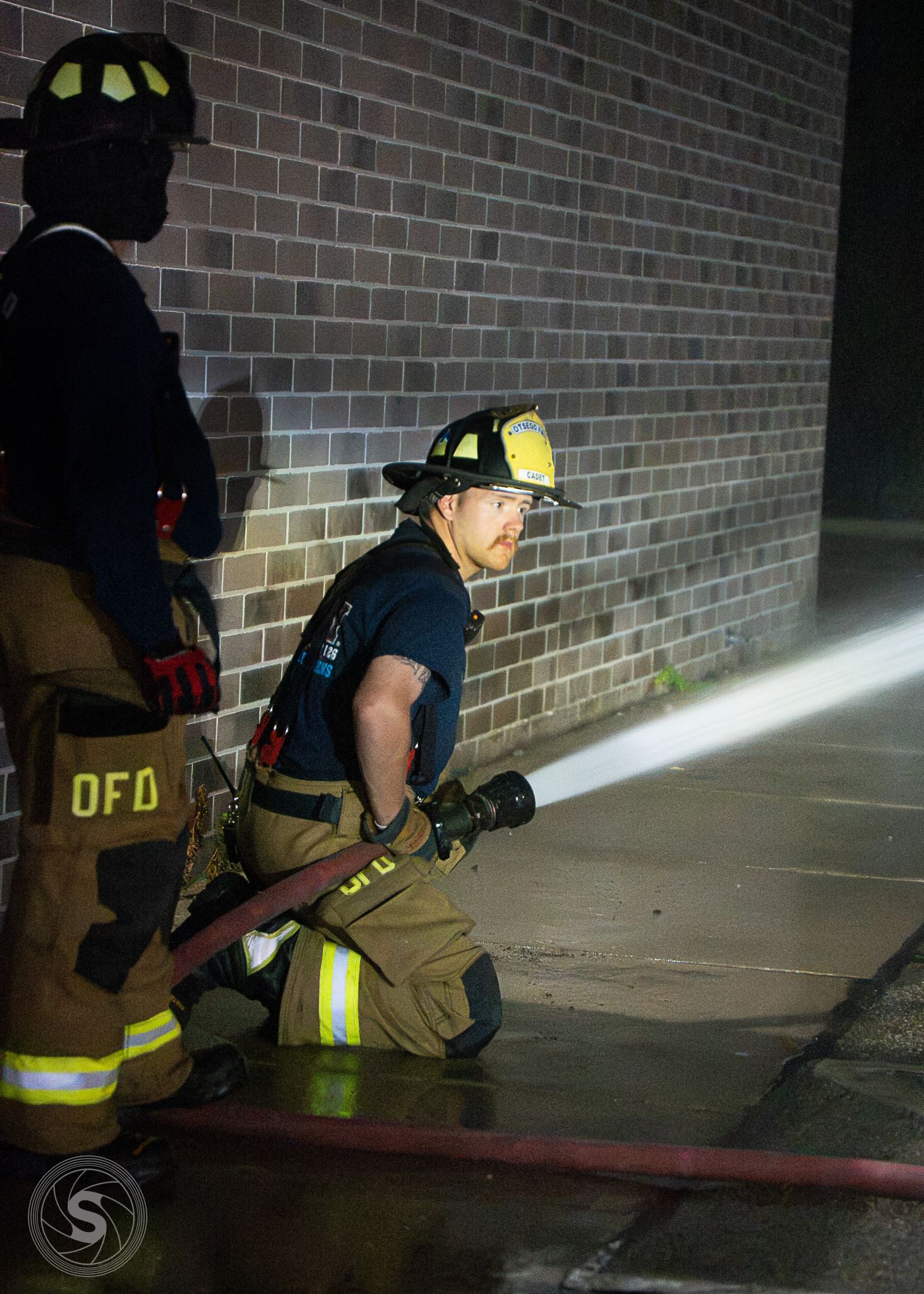 Two firefighters in full gear operate a hose outside a brick building at night, illuminated by a bright light.
