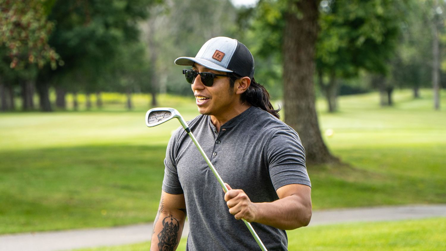 Smiling man with long hair wearing sunglasses and a cap holds a golf club on a lush green golf course.