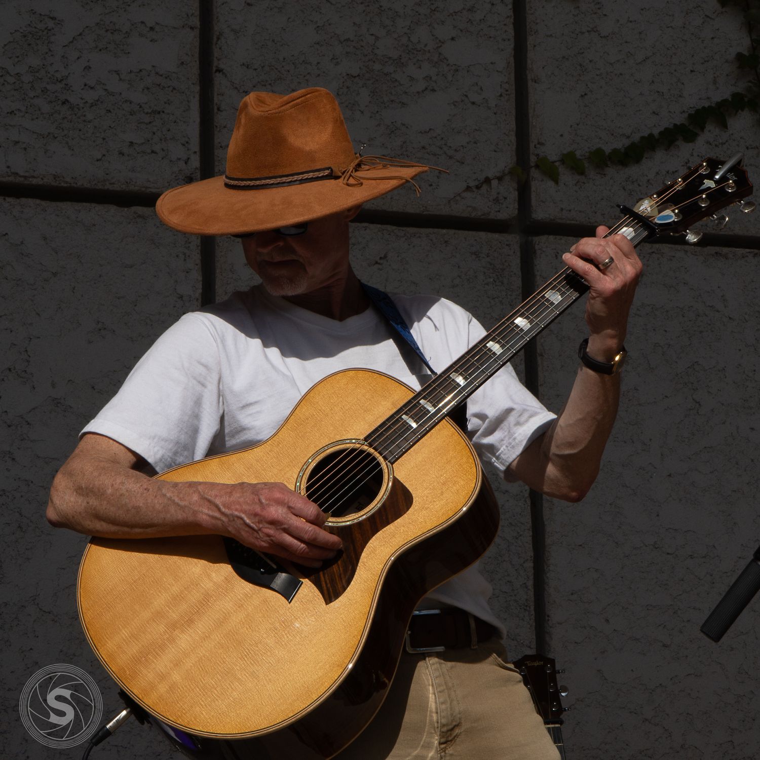 Man in brown cowboy hat playing acoustic guitar outdoors in white t-shirt at a live music event.