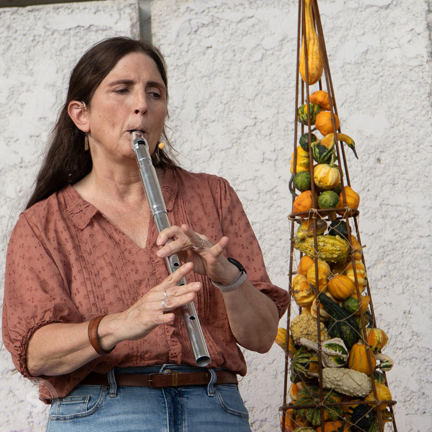 Woman playing a silver flute outdoors, wearing a pink blouse, beside a decorative pyramid of colorful gourds.