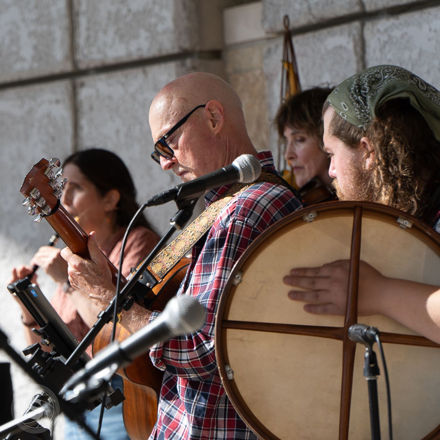 Folk musicians perform outdoors, playing guitar, violin, and drum at a live acoustic music event.