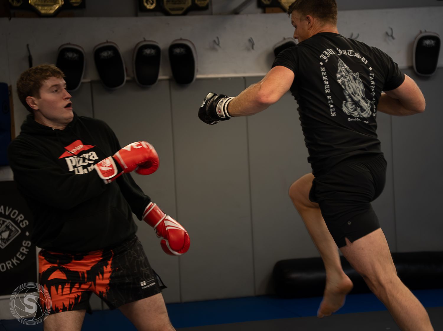 Two MMA fighters sparring in a gym, one throwing a high kick while the other defends wearing red boxing gloves.