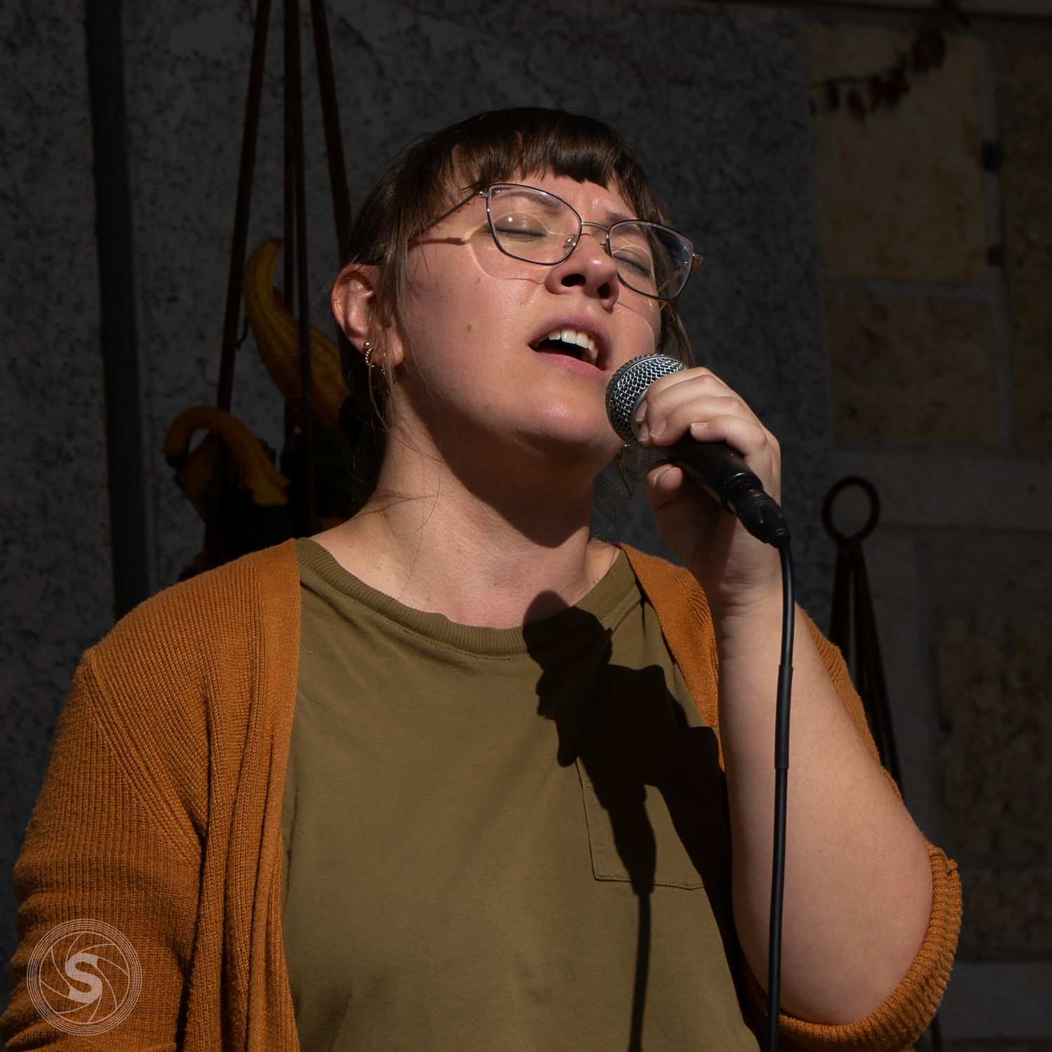 Young woman with glasses singing into a microphone outdoors, wearing a mustard cardigan over an olive green shirt.