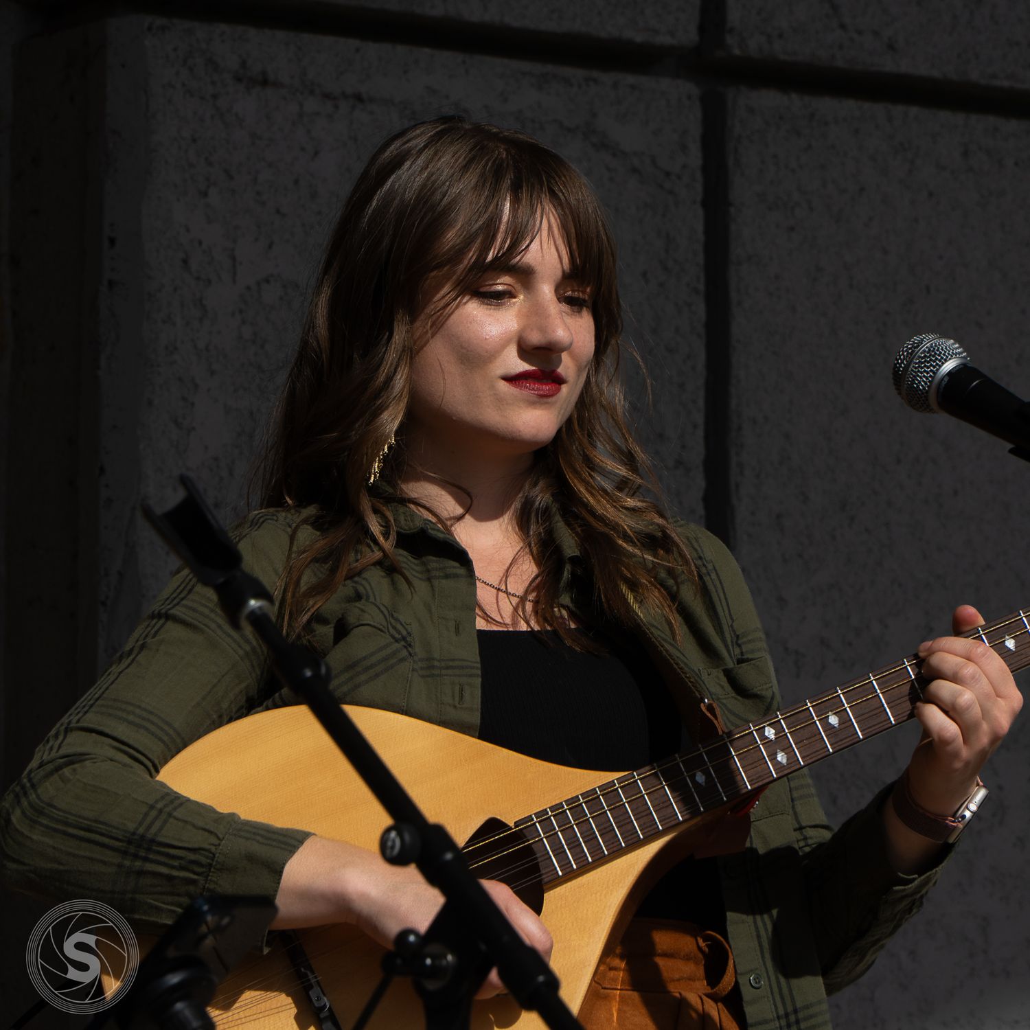 Young woman with bangs plays acoustic guitar at outdoor performance near microphone, wearing green jacket.