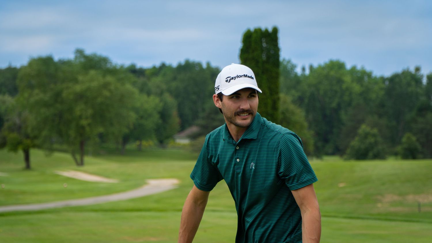 Smiling man in green polo shirt and white TaylorMade cap on a lush golf course.