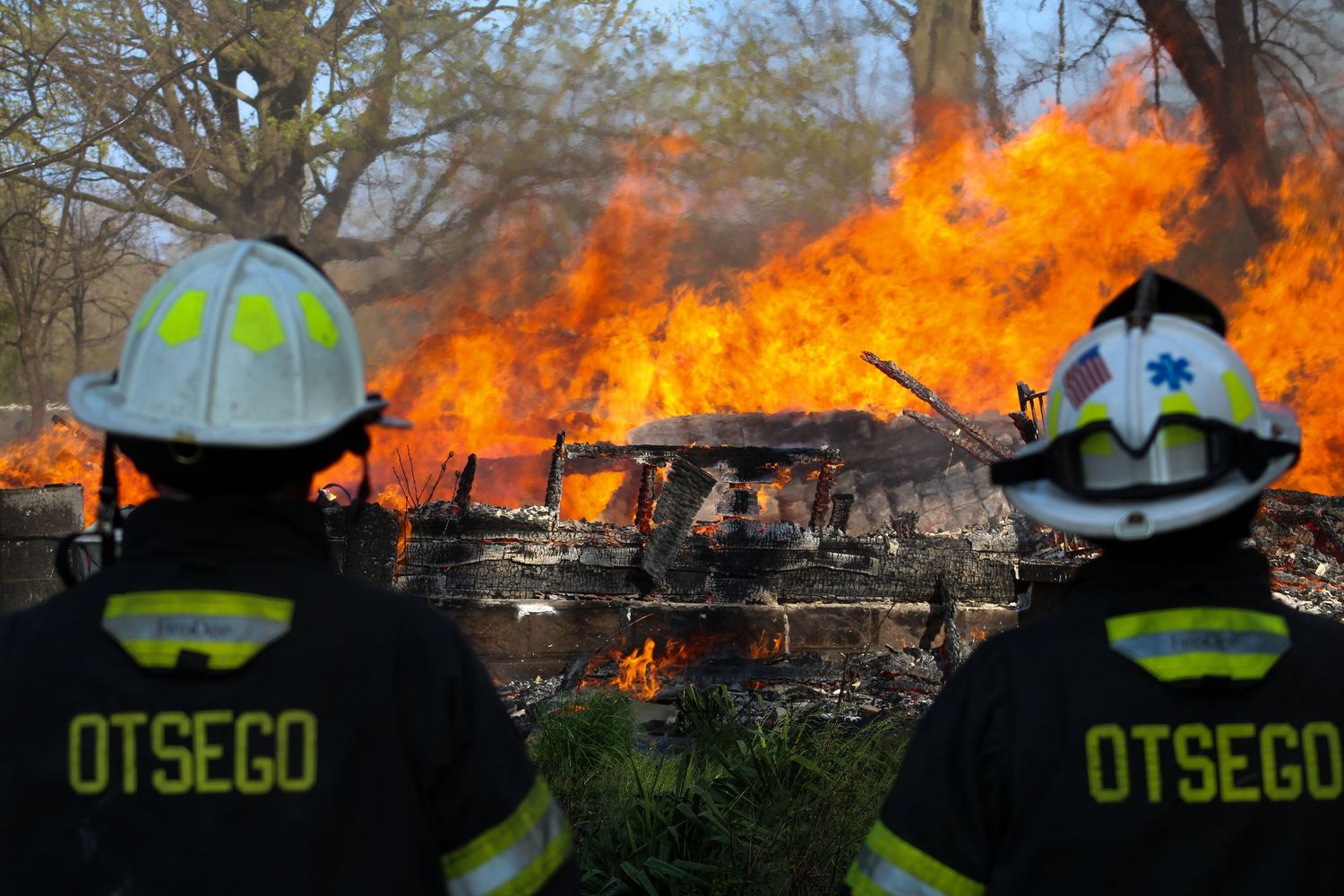 Two Otsego firefighters watch a large structure fully engulfed in flames with trees in the background.