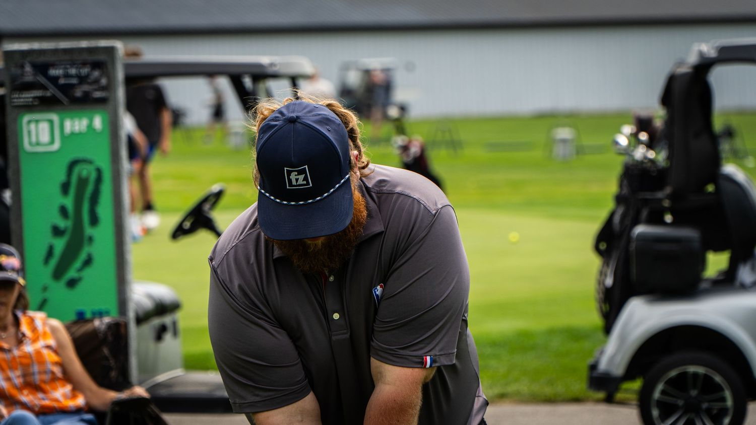 Golfer in blue cap and gray polo shirt bent over at a golf course driving range with golf carts nearby.