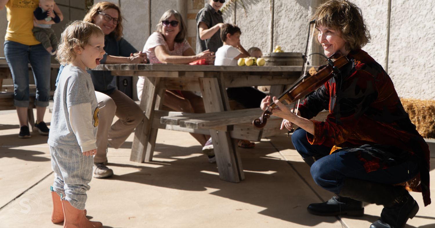 A woman plays violin for a young child outdoors at a picnic area, with adults seated at a wooden table behind them.
