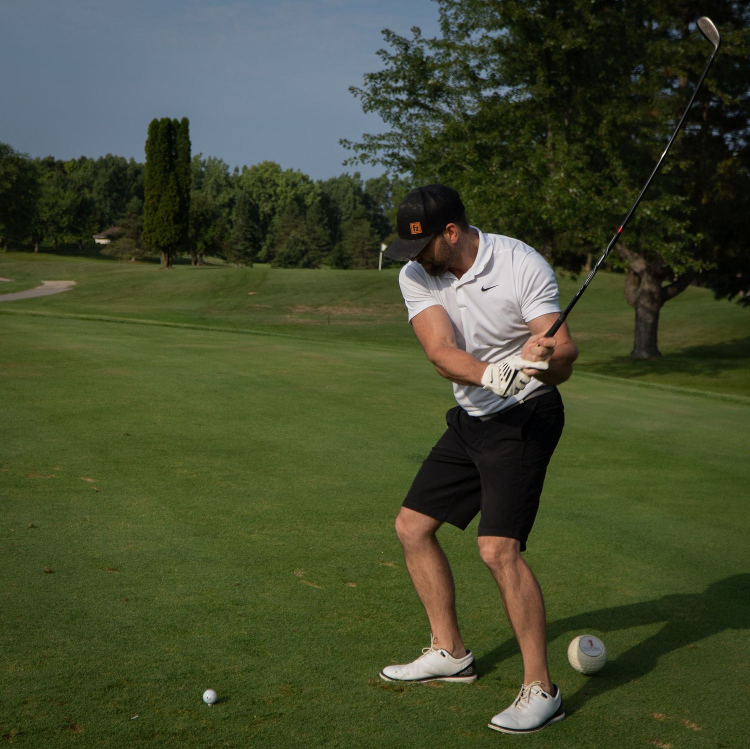 Golfer in white Nike shirt and black shorts swinging a golf club on a lush green golf course.