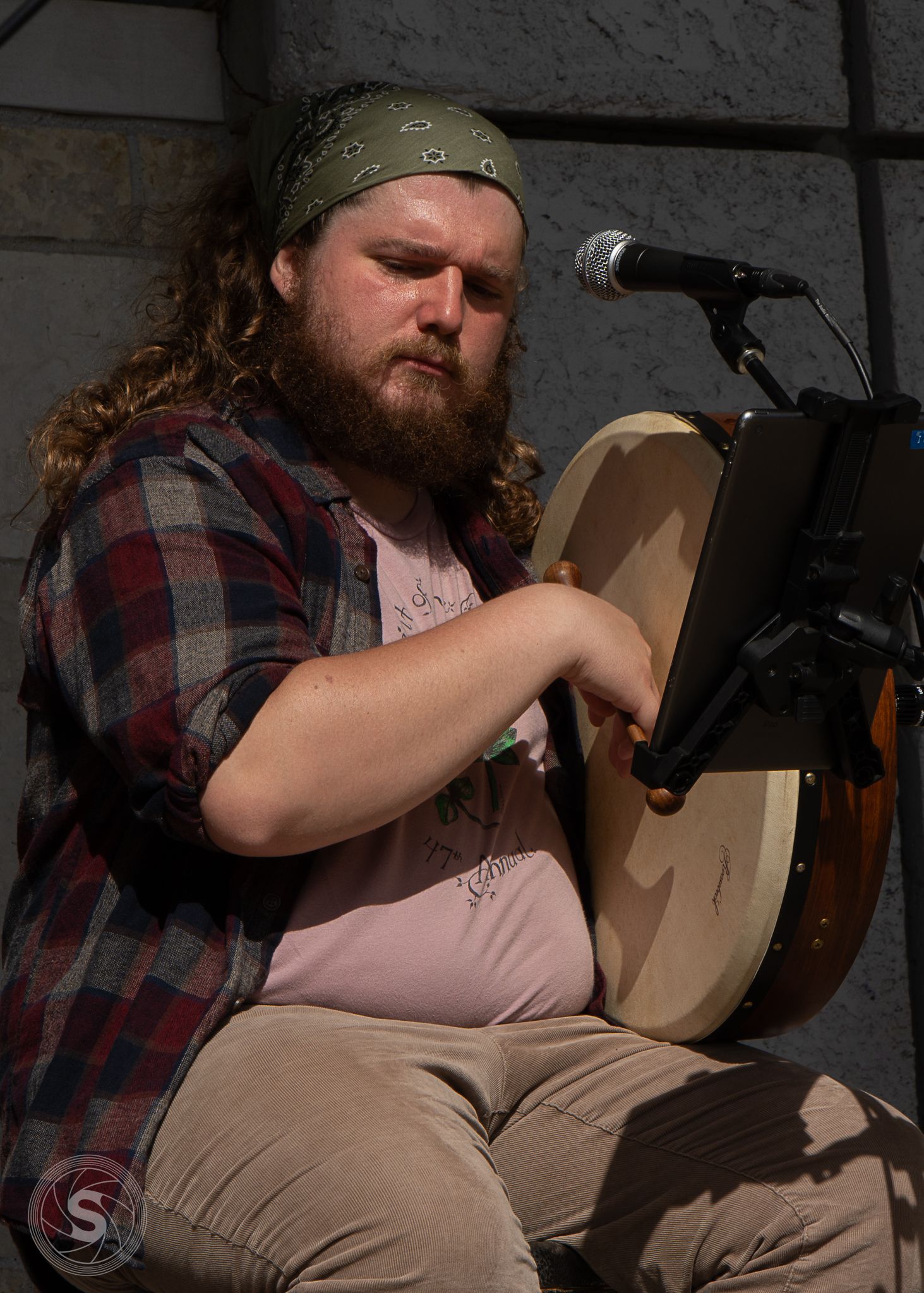 Bearded musician with long hair and green bandana plays a bodhran drum on stage near a microphone.