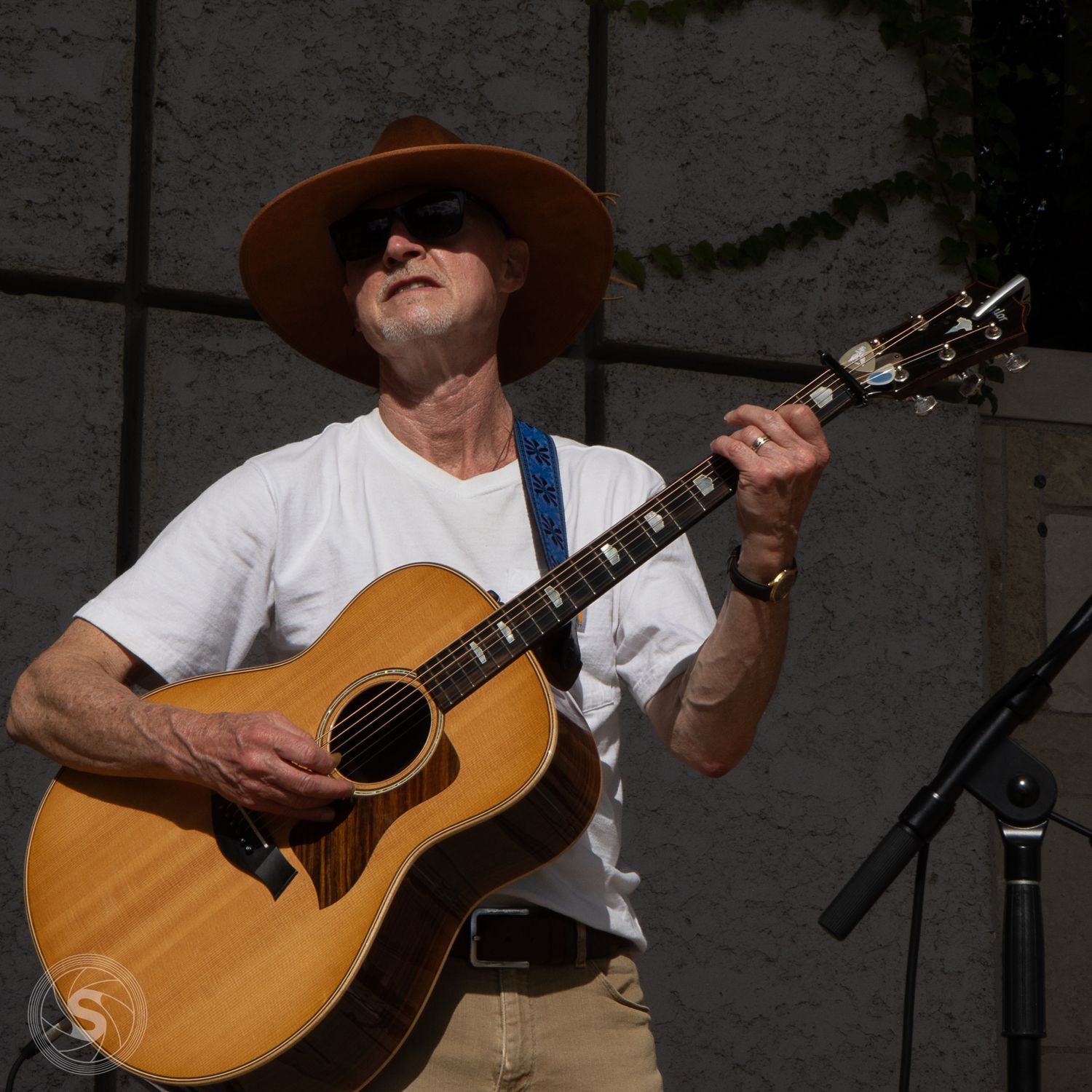 Older man wearing a brown hat and white t-shirt playing an acoustic guitar outdoors at a live music event.