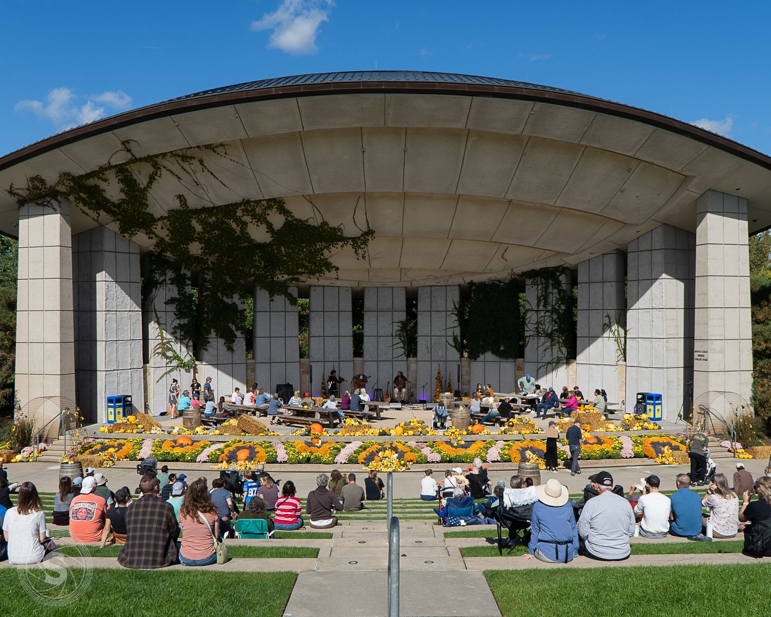Outdoor amphitheater filled with audience watching a live performance surrounded by colorful flowers on a sunny day.