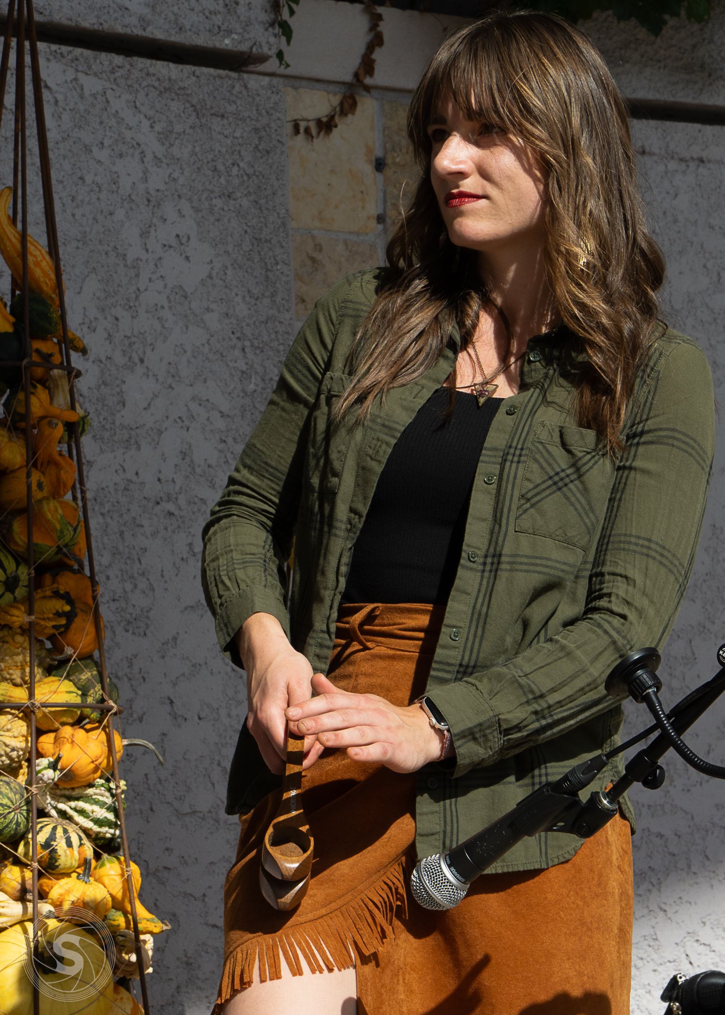 Woman in green plaid shirt and brown fringe skirt stands near autumn gourds display with microphone nearby.