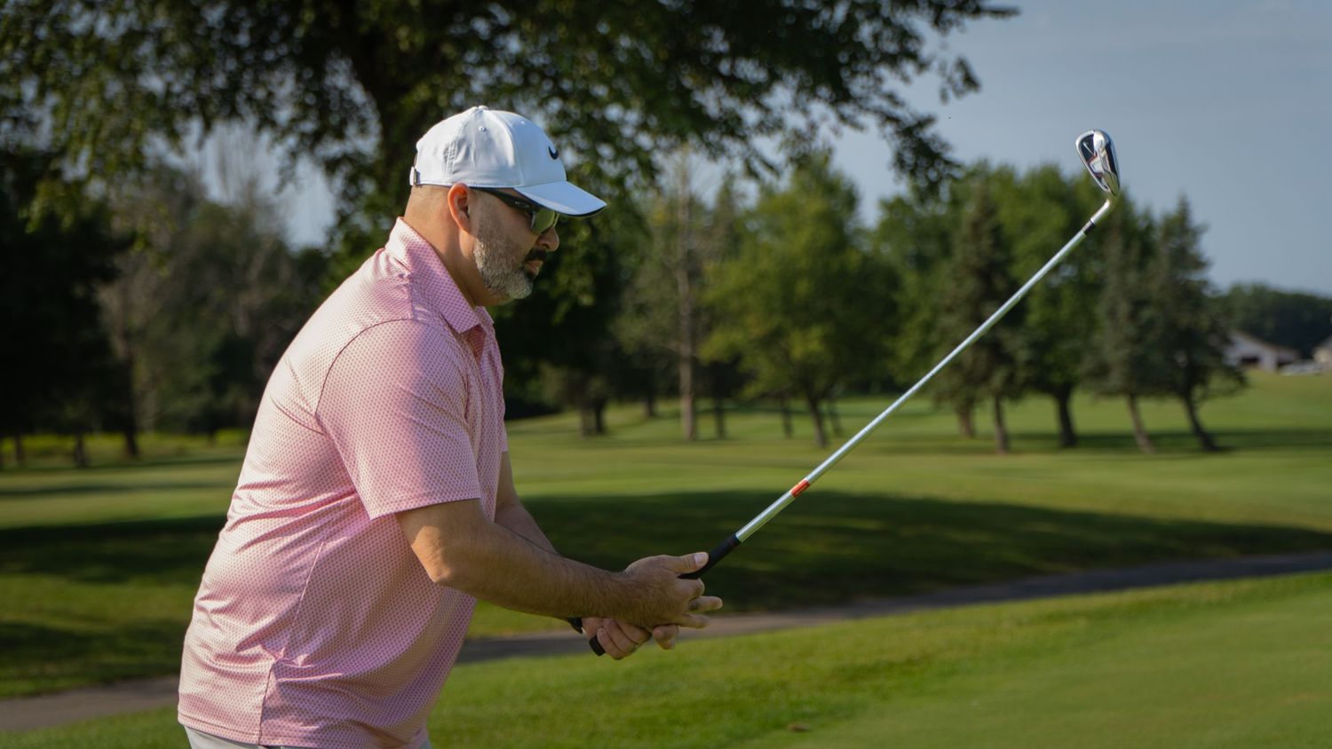 Man in pink shirt and white cap swinging a golf club on a sunny green golf course.