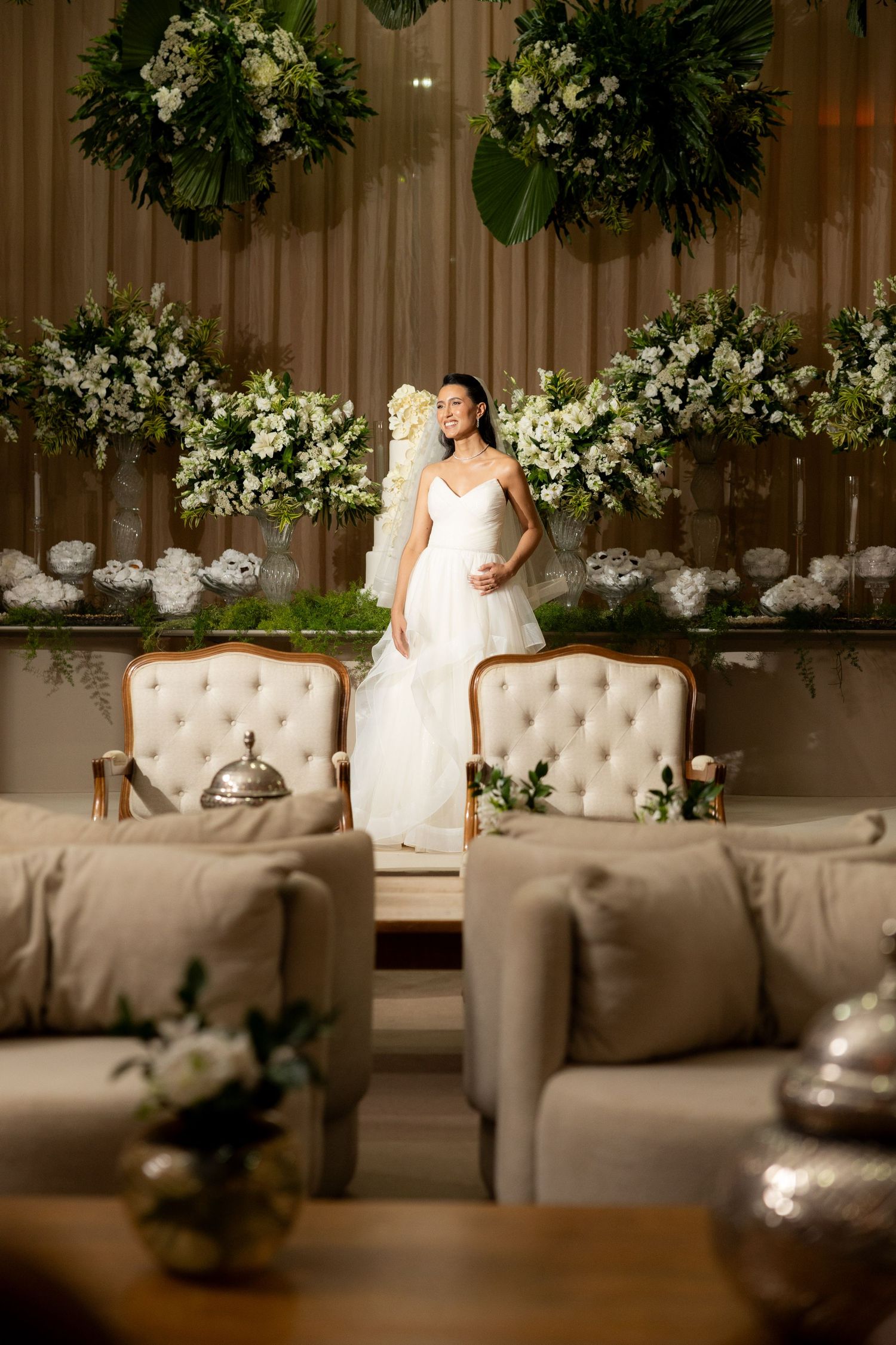 Elegant wedding venue setup with white floral arrangements and tufted seating against draped backdrop.