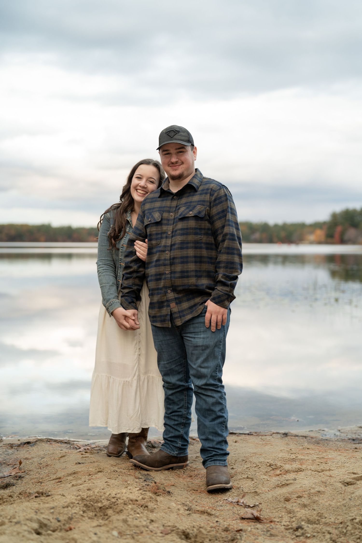 Couple stands together on sandy beach with lake and cloudy sky in background.