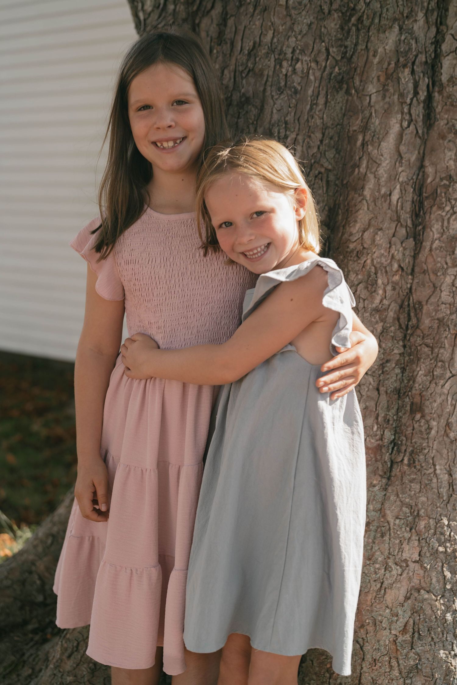Two children in pink and gray dresses hugging against tree trunk during photo session.