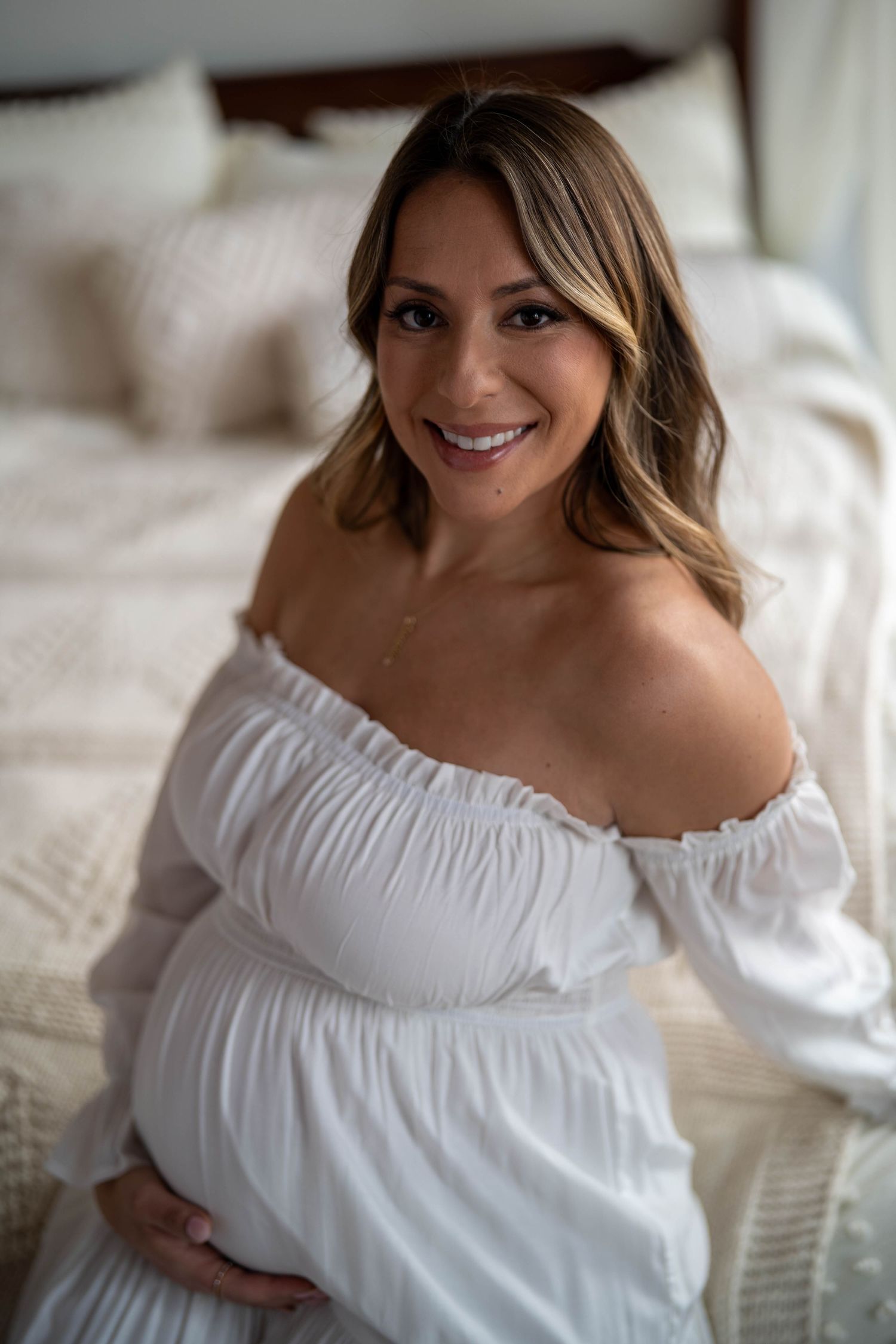 Intimate maternity portrait in white off-shoulder dress on bed.