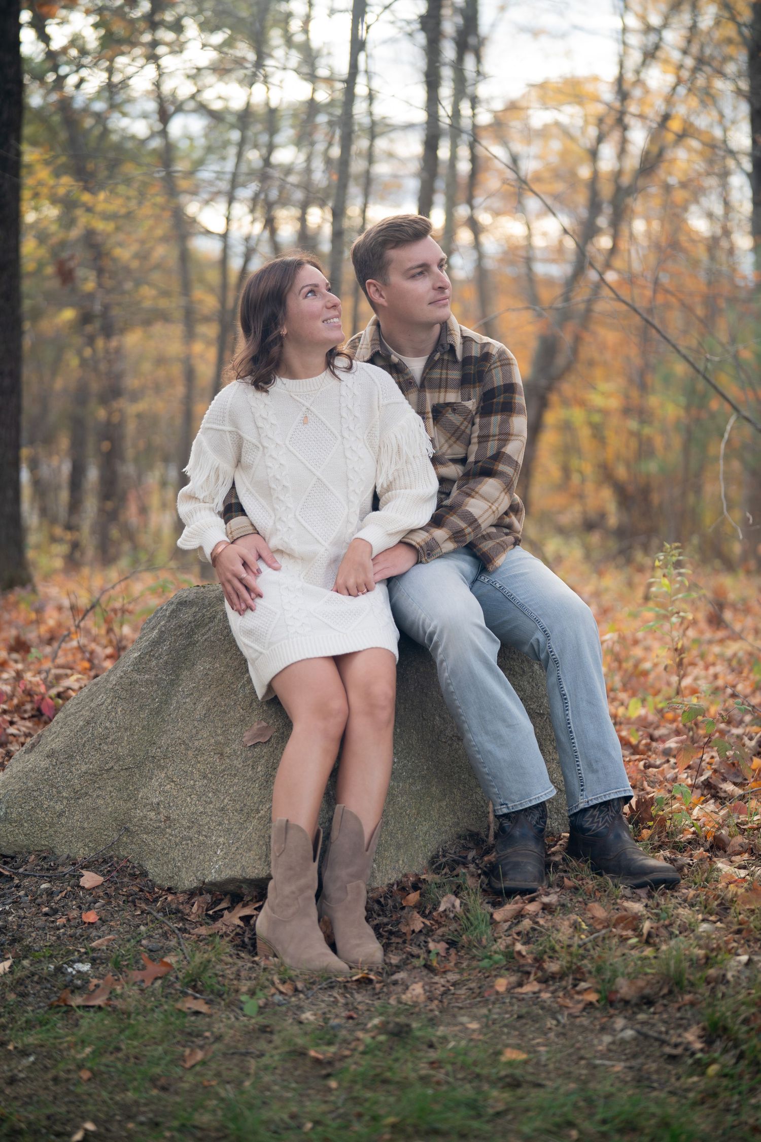 A couple sits together on a large rock surrounded by fallen autumn leaves in a wooded setting.