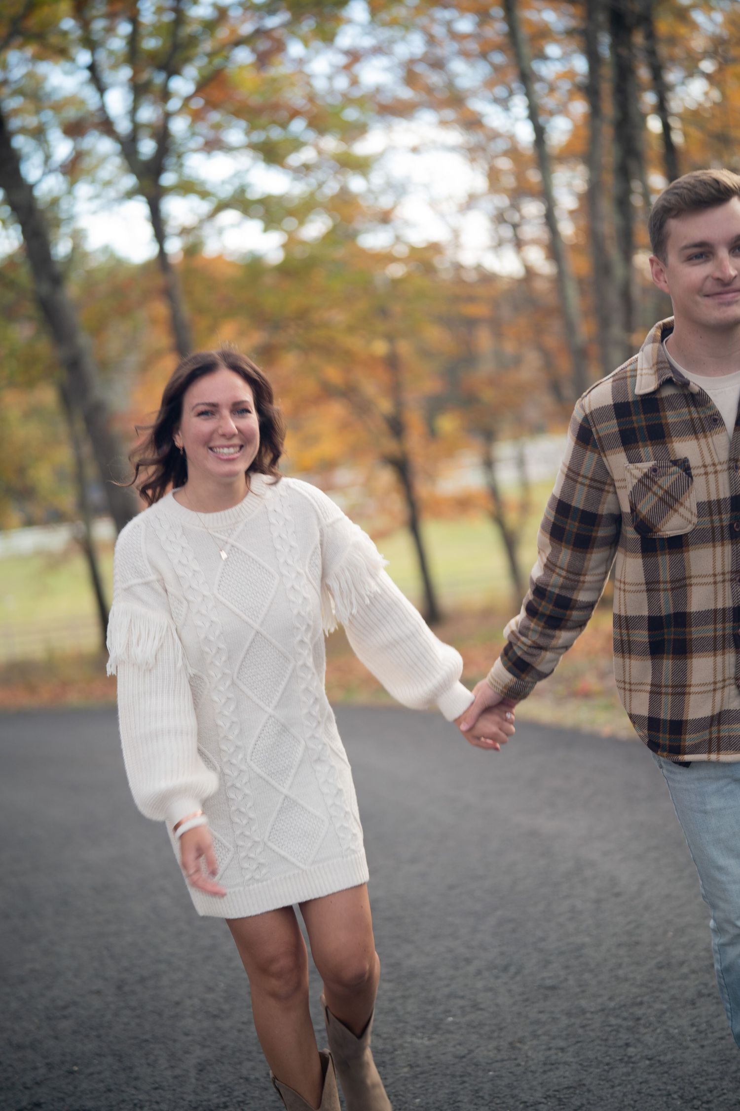 A couple walks together in a park during autumn, wearing a white sweater and plaid shirt against a backdrop of fall trees.