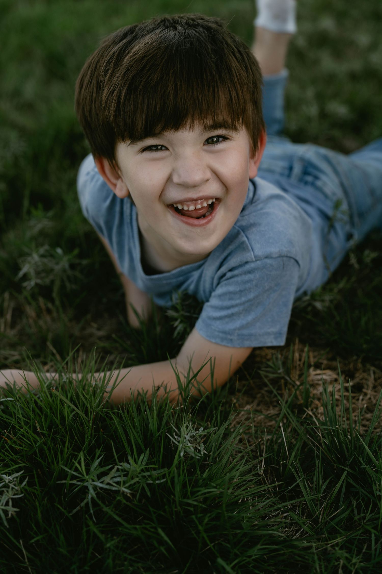 A child wearing a blue shirt lies in lush green grass while smiling at the camera.