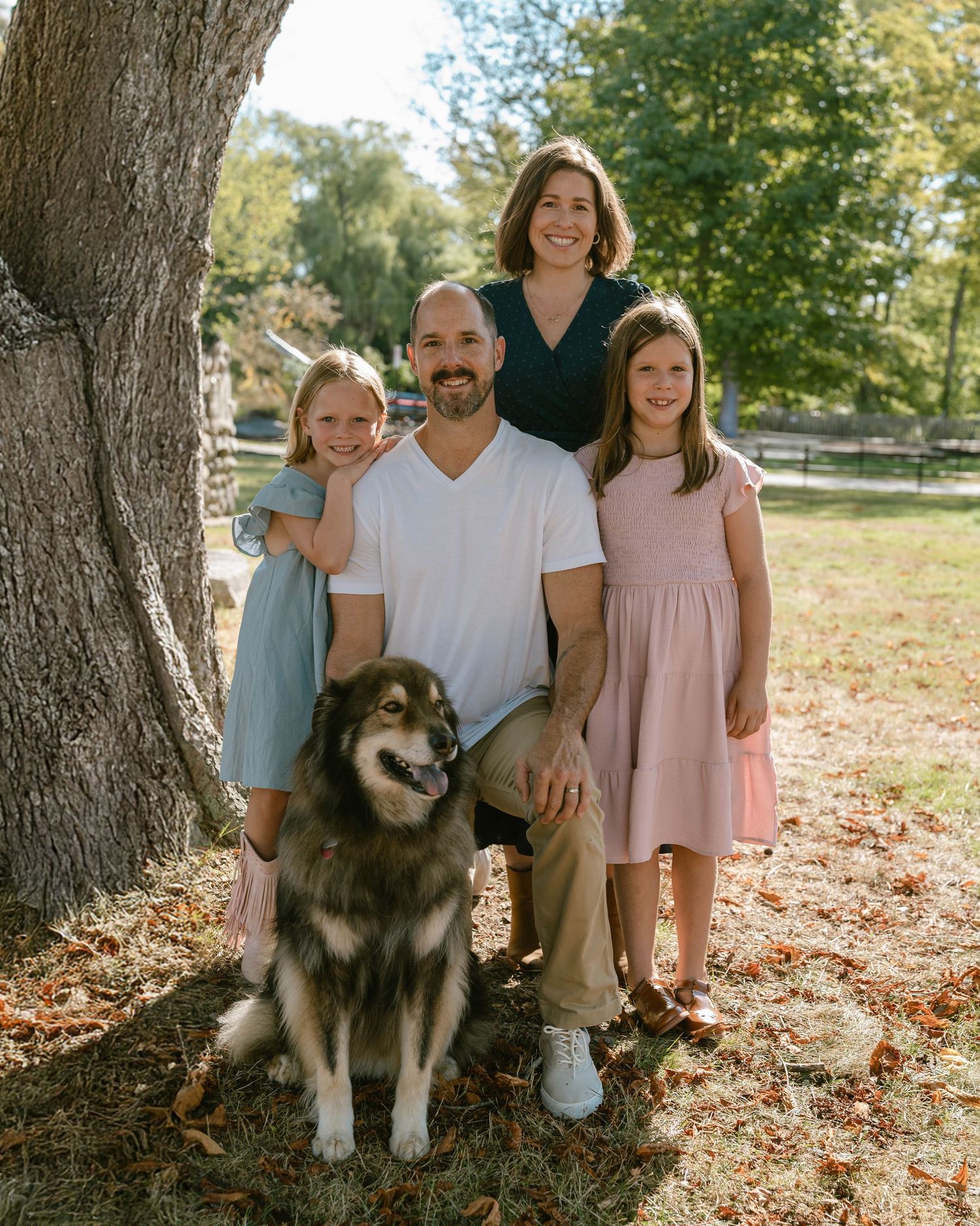 Family photo session under large tree with fluffy dog in autumn setting.